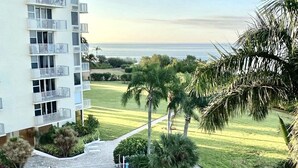 A pool area with a view of the ocean, surrounded by palm trees and a building with balconies. - ELEVATE IN THE SUN! A Renovated Beachfront Condo! (Fort Myers Beach)