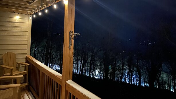 A wooden deck with a railing, string lights, and a view of a forested area at night. - APPresSki - 5 bed Ski in Ski out - 50 Steps to Ski Sugar Mountain Slopes (Sugar Mountain)