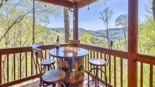 A wooden deck with a round table, bar stool, and a bottle of wine. - Long- & Short-Range Views/King Beds/Fireplace/Fire Table/Game Room (Maggie Valley)