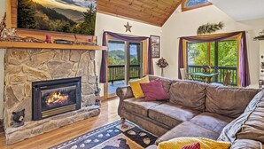 A cozy living room with a stone fireplace, a leather sofa, and a large window overlooking a deck. - Long- & Short-Range Views/King Beds/Fireplace/Fire Table/Game Room (Maggie Valley)