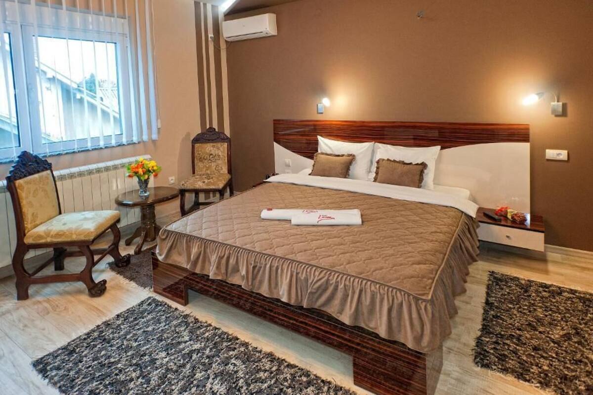 A hotel room with a large bed, a wooden nightstand, a chair, a small table with a vase of flowers, and a window with blinds.