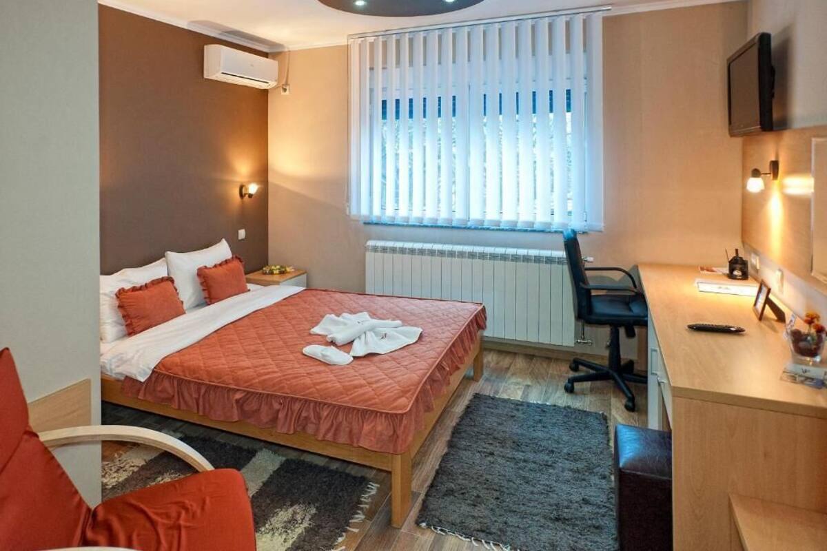A hotel room with a bed, a desk, a chair, a TV, and a window with blinds.