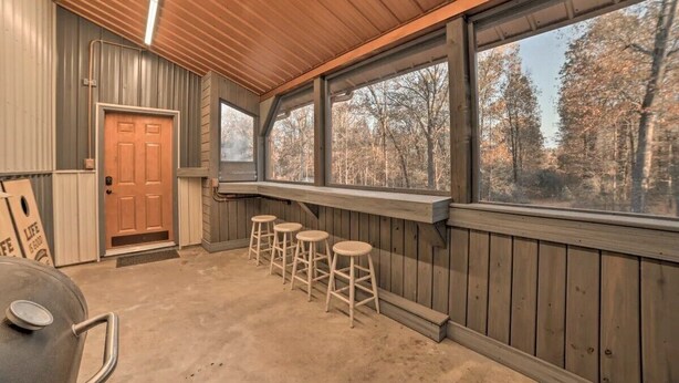 A covered outdoor area with a wooden ceiling, a door, and large windows overlooking a forest. There are four wooden stools and a barbecue grill.