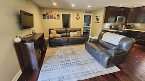A living room with a dark brown leather sofa, a flat-screen TV, a patterned rug, and a kitchen area with dark wooden cabinets and a microwave.