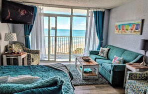 A room with a bed, a sofa, a TV, a table, and a view of the beach.