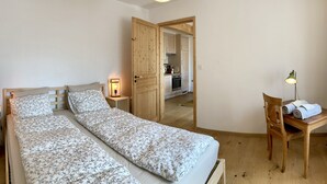 A room with two beds, a desk, a chair, a lamp, and a door leading to a kitchen. - Apartment (Fontainemelon)