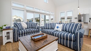 A spacious living room with a large window, a striped sofa set, a wooden coffee table, and a kitchen area with white cabinets. - Upscale Family Home for 10, Community Pool, Grill (Ocean City)