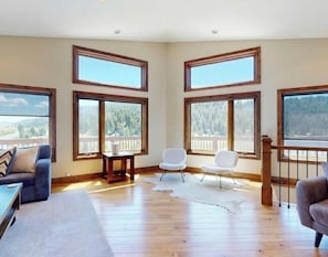 Una habitación amplia con ventanas grandes, suelo de madera, alfombra blanca y vistas a un área boscosa.