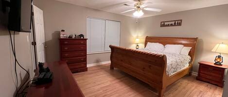 A bedroom with a wooden bed, a dresser, a desk, a ceiling fan, and a television mounted on the wall.