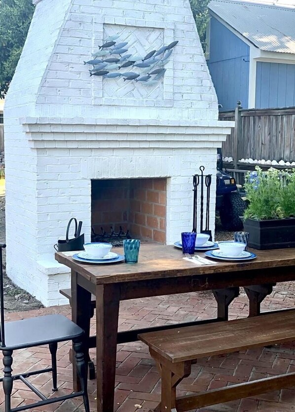 Un horno de ladrillo exterior con revestimiento blanco y una chimenea, rodeado por una mesa de madera con platos, cuencos y cubiertos.