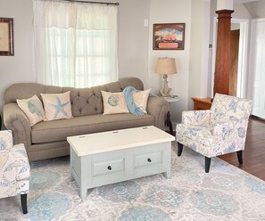 Una sala de estar con un sofá beige, un sillón estampado y una mesita de noche blanca con una lámpara.