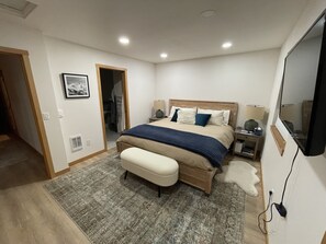 Un dormitorio con cama, mesitas de noche, un banco, una alfombra, un cuadro en la pared y un espejo.