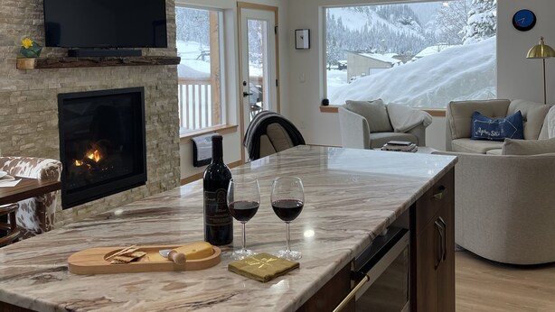 Una cocina moderna con pared de piedra, chimenea, una isla grande con copas de vino y vistas a un paisaje nevado.
