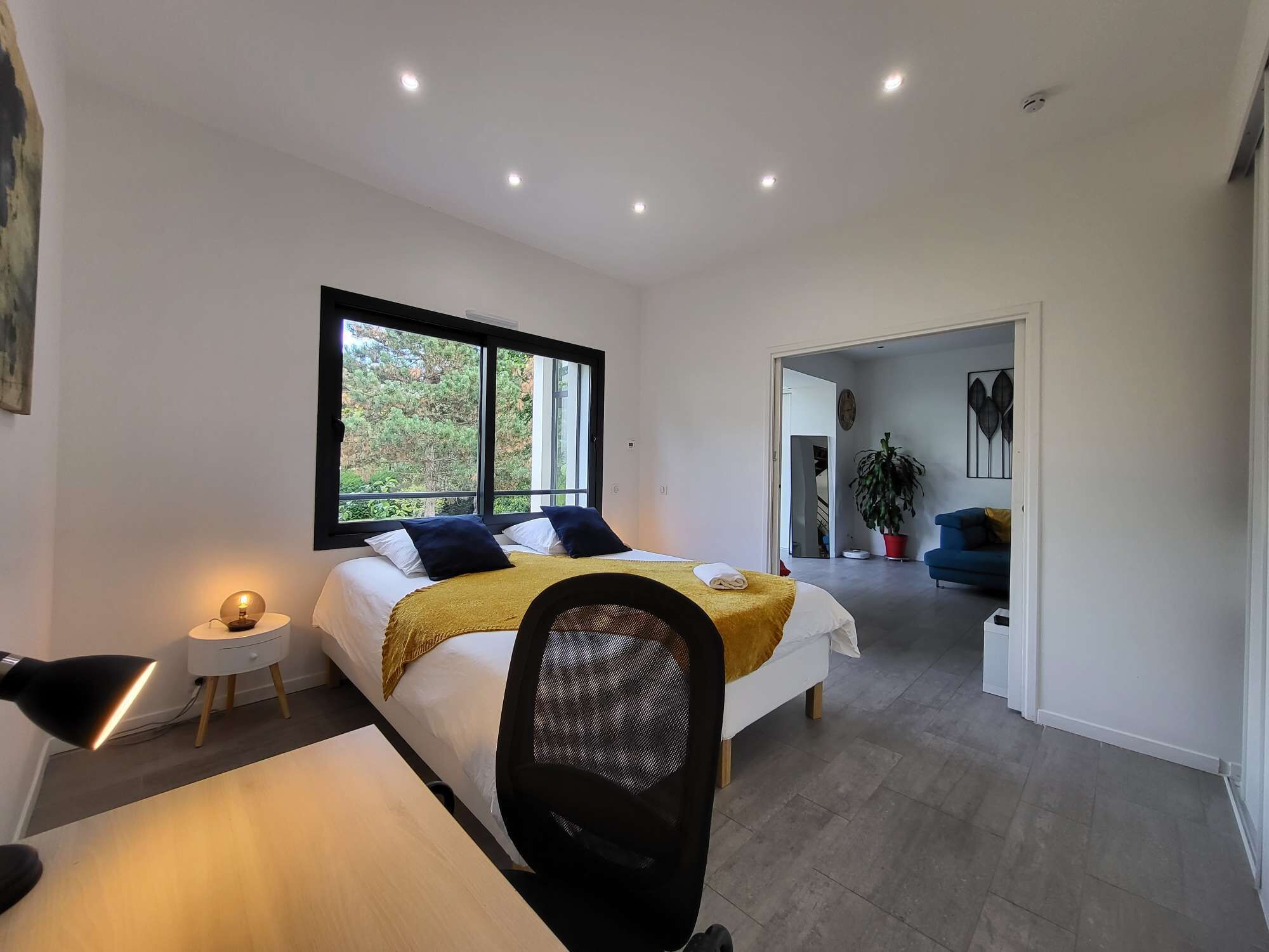 Une chambre moderne avec un grand lit, un bureau et une chaise. Une fenêtre offre une vue sur les arbres à l’extérieur.