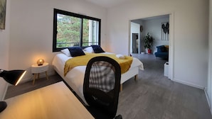 Un dormitorio moderno con una cama grande, un escritorio y una silla. Se ve una ventana con vistas a árboles.