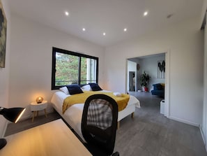 Une chambre moderne avec un grand lit, un bureau et une chaise. Une fenêtre offre une vue sur les arbres à l’extérieur.