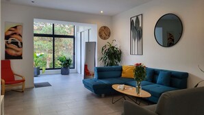 Un salón moderno con un sofá azul, un sillón rojo, una mesa de centro y grandes ventanales con vistas al verdor.