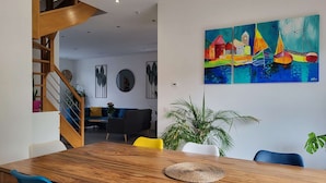 Un comedor con una mesa de madera, sillas amarillas y azules, una escalera y un cuadro colorido en la pared.