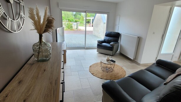A modern living room with a wooden sideboard, a black leather sofa, a round coffee table, and a large window with a view of the outdoors. - House located in a quiet cul-de-sac, close to the dunes. (Créances)
