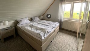 A bedroom with a sloped ceiling, a bed with star-patterned bedding, a nightstand, a window with a view of greenery, and a radiator. - Vacation apartment Assing am Deich (Dornum)