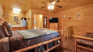 Ein gemütlicher Holzblockraum mit Bett, Fernseher, einer kleinen Küchenzeile und einem Deckenventilator.