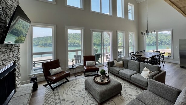 A spacious living room with large windows, a stone fireplace, and a dining area with a view of the lake. - Modern Lake Cottage - Amazing Views - 6 Bed/6 Bath - Boat Launch (LaFollette)