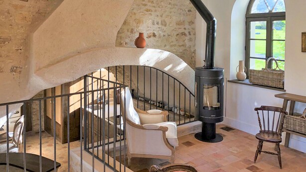 A rustic interior with a stone wall, a black wood-burning stove, a white chair, and a wicker basket by a window.