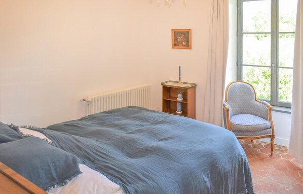 Un dormitorio con cama, una silla, un radiador, un cuadro en la pared y una ventana con vista a follaje.