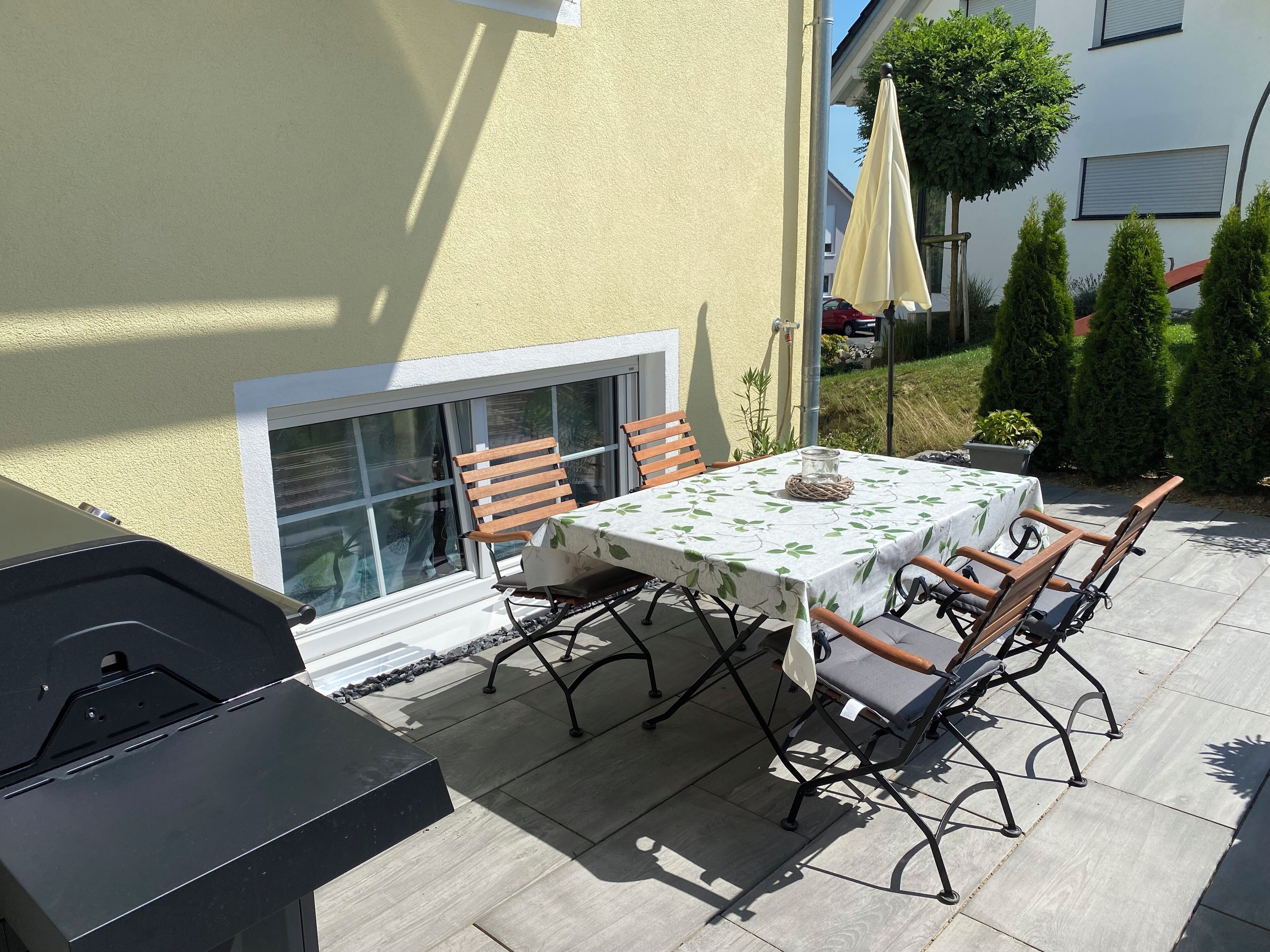 Une terrasse extérieure avec une table dressée pour deux personnes, entourée de chaises, d’un barbecue et d’une plante en pot.