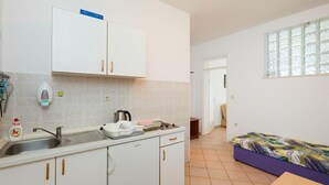 Une cuisine compacte avec des meubles blancs, un évier et une bouilloire. On aperçoit un lit avec une couvre-lit colorée dans la pièce adjacente.