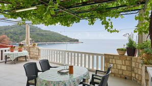 Une terrasse avec vue sur la mer, aménagée avec des tables et des chaises, des plantes en pot et une pergola couverte de plantes grimpantes.