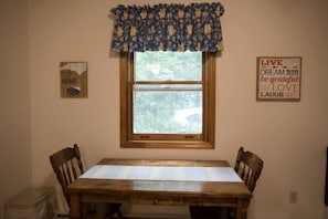 Un comedor con una mesa y sillas de madera, una ventana con un cortinero floral, y dos carteles con mensajes inspiradores en la pared.