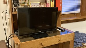 Un meuble en bois pour la télévision, sur lequel est posée une télévision Samsung, avec une télécommande et une petite étagère au-dessus, servant à ranger des livres et des objets décoratifs.
