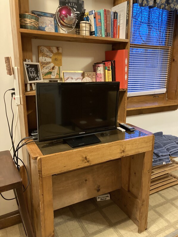 A wooden TV stand with a Samsung TV, a remote control, and a small shelf above holding books and decorative items.