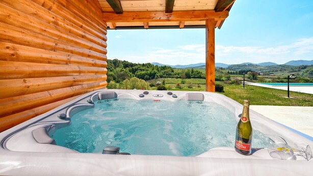 Un jacuzzi con una botella de vino y copas, frente a un paisaje con colinas y un lago.