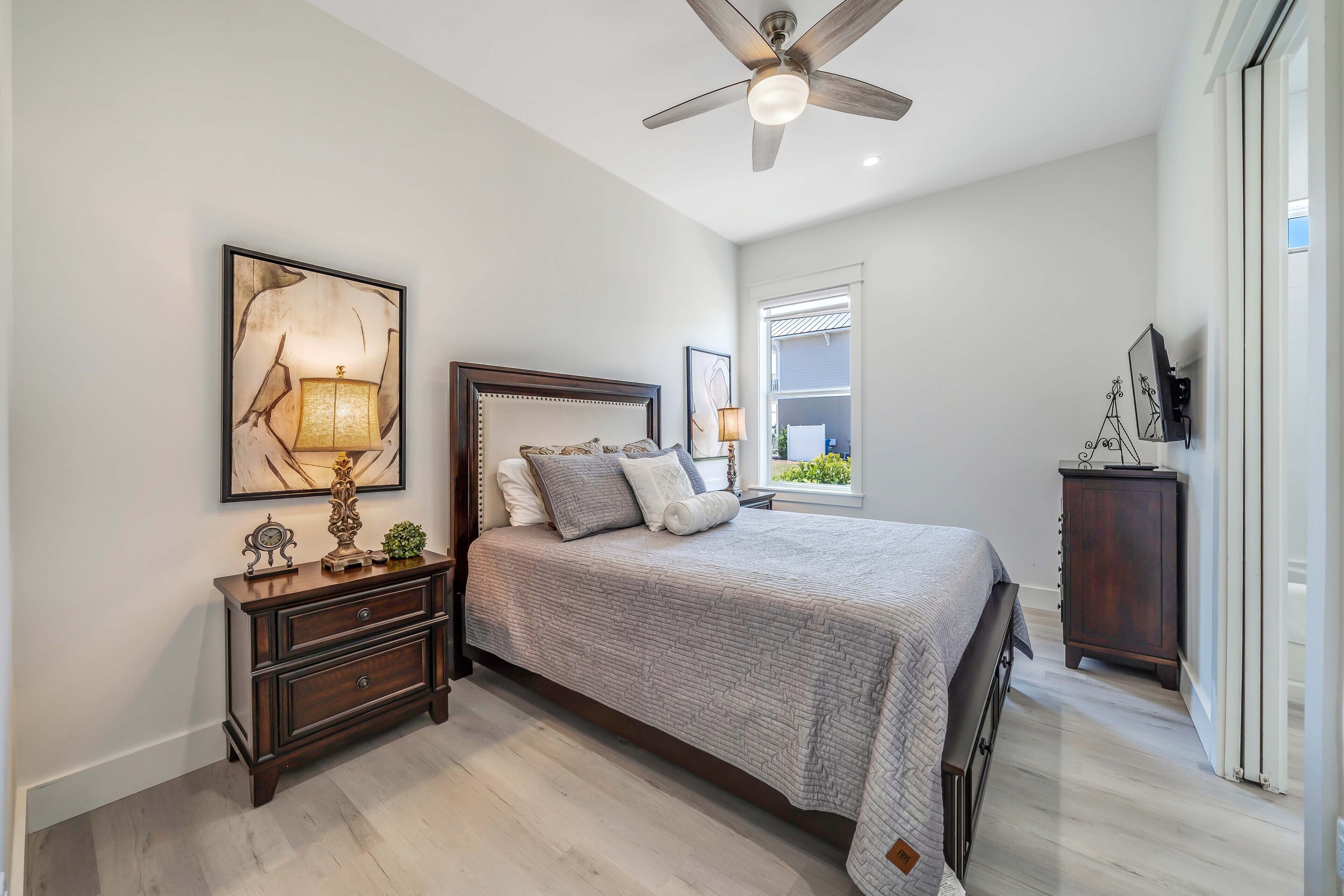 A bedroom with a bed, nightstand, wall art, ceiling fan, and a window.