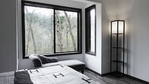 Une chambre avec une grande fenêtre, une table de chevet, une chaise et une lampe sur pied.