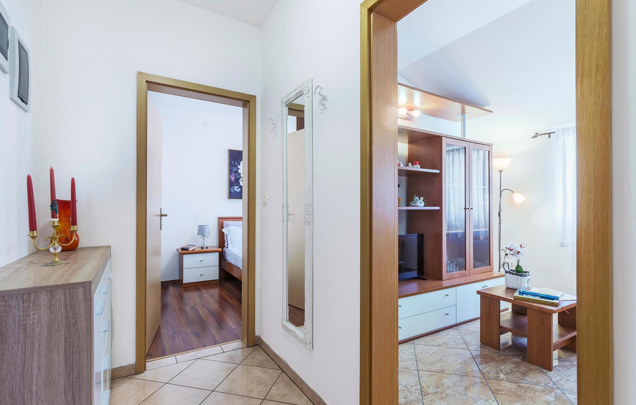 A hotel room with a wooden wardrobe, a bedside table, a desk with a chair, and a mirror.