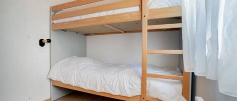 Una cama individual con estructura de madera y ropa de cama blanca en una habitación con suelo de madera y paredes blancas.