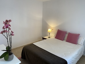 A neatly made bed with a white and brown bedspread, two pink pillows, a bedside lamp, and a potted orchid plant on a white table. - Padrón Centro, Camino De Santiago (Padrón)