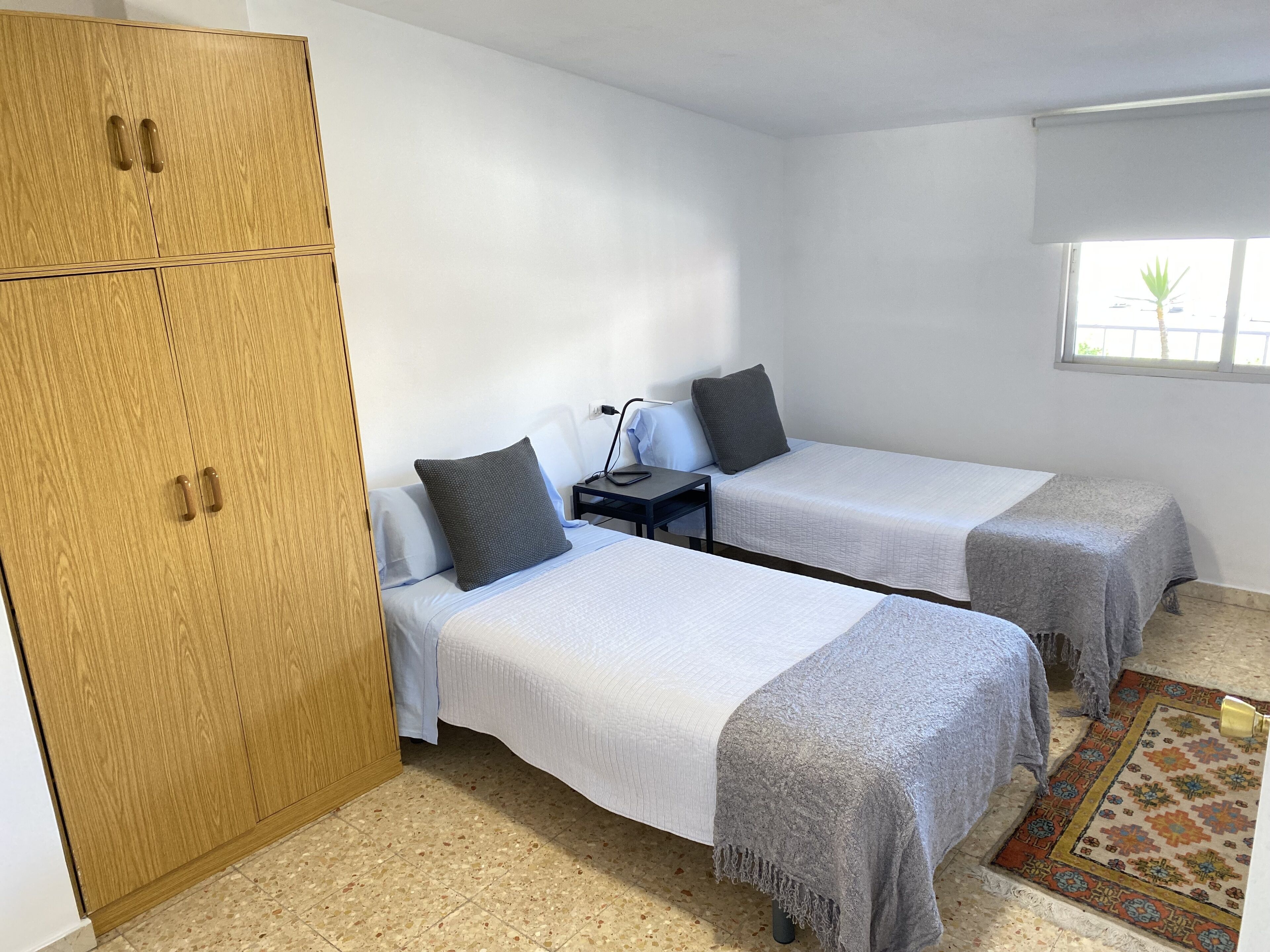 Habitación con dos camas individuales, un armario de madera, una mesita de noche con lámpara y ventana con vistas a zonas verdes.