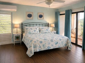 Ein Schlafzimmer mit einem Bett, zwei Nachttischen, einem Deckenventilator, einem Fenster mit Vorhängen und Blick ins Freie.