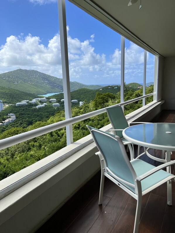 Ein Balkon mit Glastisch und Stühlen, der auf eine üppige grüne Landschaft und eine entfernte Ansicht von Gebäuden und Hügeln blickt.