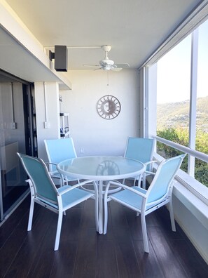 Ein Balkon mit rundem Glastisch und vier Stühlen, einem Deckenventilator und einem an der Wand befestigten Dekorationsgegenstand.
