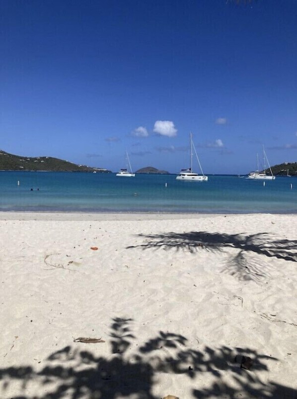 A beach with white sand and clear blue water. There are several sailboats anchored in the distance.