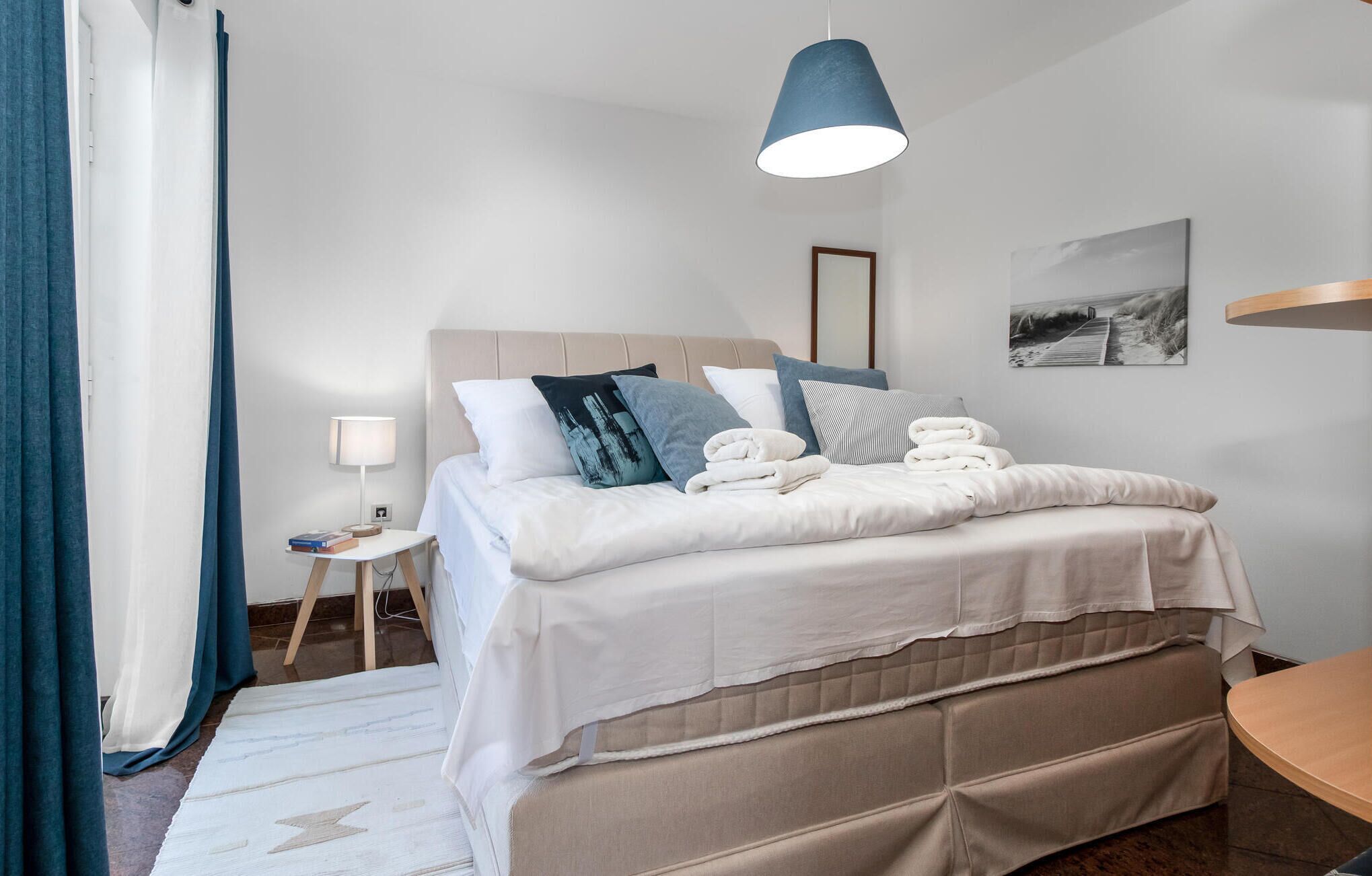 A neatly made bed with multiple pillows and folded towels, a bedside table with a lamp, and a hanging light fixture above the bed.