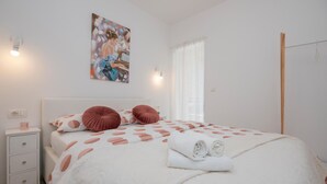 Ein ordentlich eingerichtetes Schlafzimmer mit Bett, Kissen, Nachttisch, einem Gemälde und einem Fenster mit Vorhängen.