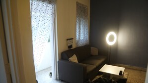 Una habitación pequeña, con poca luz, que tiene un sofá gris, una mesita de noche blanca y un aro de luz.