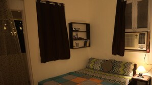 Un dormitorio pequeño y acogedor con una cama individual, una mesita de noche con lámpara, una ventana con aire acondicionado, un estante con libros y una cortina.