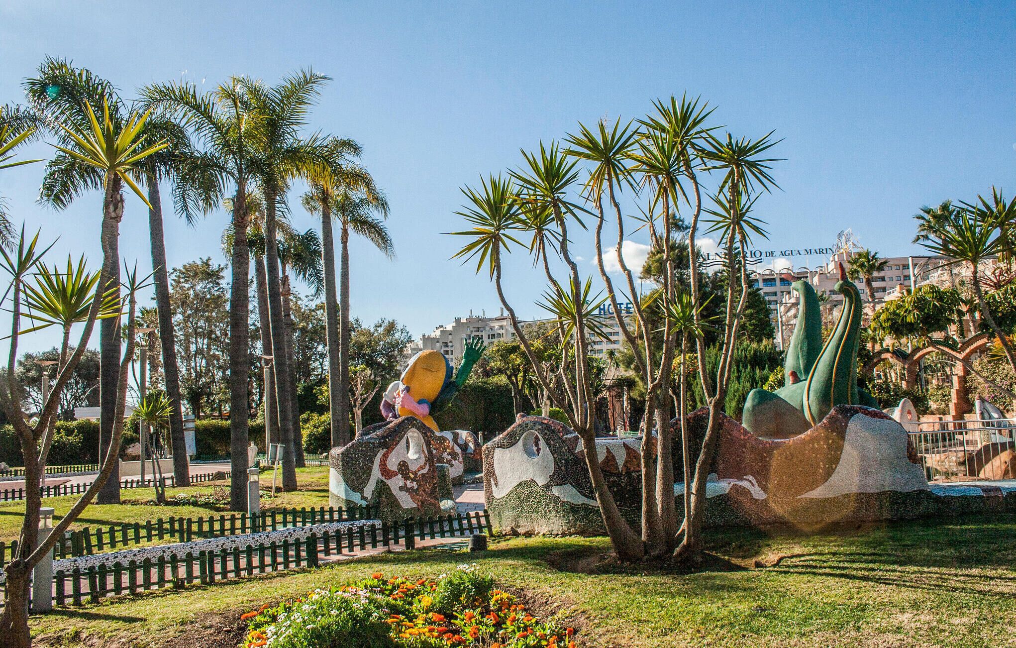 Un parc avec des palmiers, une fontaine et un bâtiment dont on aperçoit une enseigne au fond.